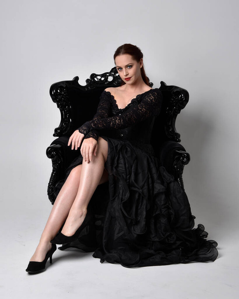 Siyahi gotik elbiseler giyen, süslü siyah bir koltukta oturan bir kadının tam boy portresi. Oturmuş poz, stüdyo arka planında.. - Fotoğraf, Görsel
