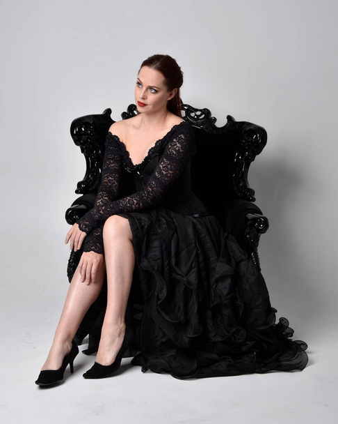 full length portrait of  woman wearing black gothic dress, sitting on a ornate black armchair. Seated pose, against a studio background. - Foto, Bild