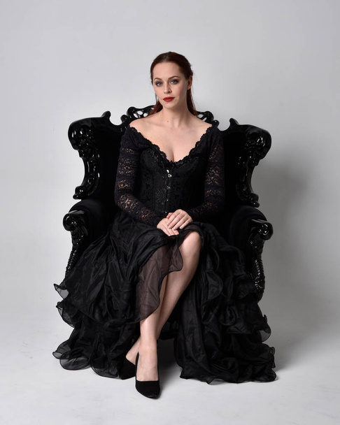 volledig portret van een vrouw in zwarte gotische jurk, zittend op een versierde zwarte fauteuil. Zittende houding, tegen een studio achtergrond. - Foto, afbeelding