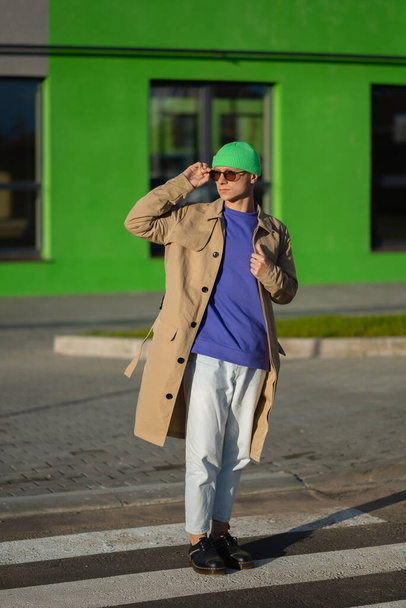 Imagen de un joven hipster feliz con ropa casual sostiene gafas de sol, aislado en el fondo de la ciudad - Foto, imagen