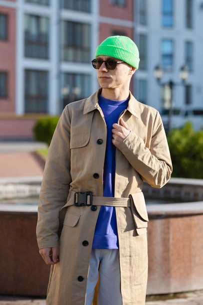 Ο νεαρός φοράει μοντέρνο παλτό, πράσινο καπέλο, λευκό τζιν και πουλόβερ. - Φωτογραφία, εικόνα