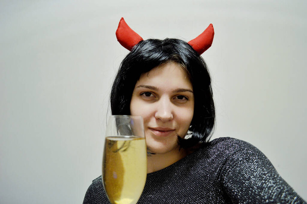 Mulher com cabelo preto curto com chifres vermelhos e uma taça de champanhe na mão sobre um fundo claro - Foto, Imagem