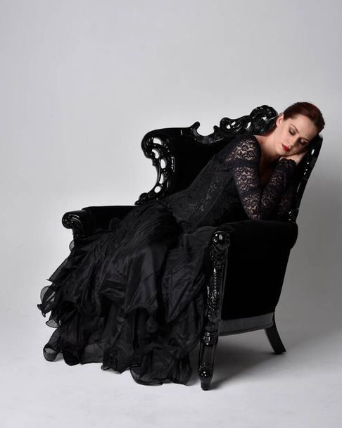 full length portrait of  woman wearing black gothic dress, sitting on a ornate black armchair. Seated pose, against a studio background. - Photo, Image