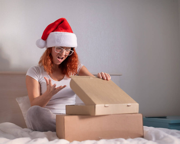 Upset woman in santa claus hat unpacks online purchase. The girl received the wrong parcel. - Foto, afbeelding