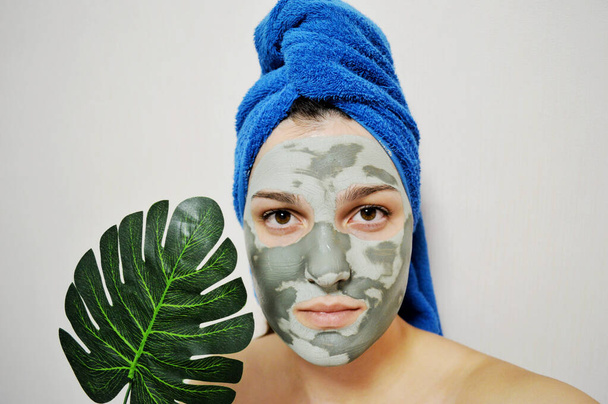 Kafasında mavi havlu ve yüzünde kilden bir maske olan kadın bir palmiye yaprağı tutuyor. - Fotoğraf, Görsel