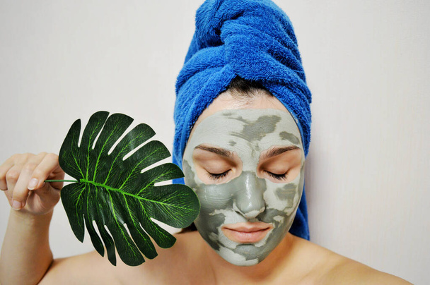 Femme avec une serviette bleue sur la tête et un masque d'argile sur son visage tient une feuille de palmier - Photo, image