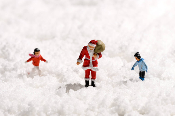Miniatuur Kerstman of Kerstman met een zak geschenken wandelen door dikke witte winter sneeuw vergezeld van een jong meisje en jongen met kopieerruimte voor een seizoensgebonden vakantie groet - Foto, afbeelding