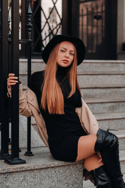 Stylish young woman in a beige coat in a black hat on a city street. Women's street fashion. Autumn clothing.Urban style. - Zdjęcie, obraz