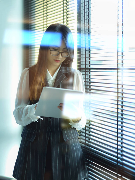 Vista a través de la ventana de vidrio de una trabajadora de oficina que trabaja con una tableta digital mientras está de pie junto a la ventana - Foto, Imagen