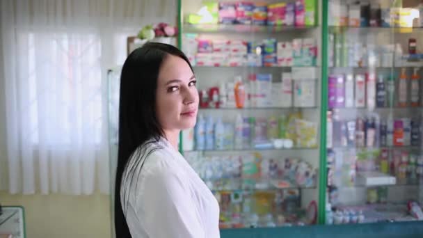 Egy gyönyörű barna nő, aki gyógyszerészként dolgozik, és egy gyógyszertár ablakának hátterében pózol. - Felvétel, videó