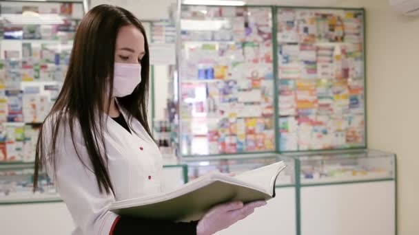 Egy nő orvosi maszkban, köntösben és kesztyűben, papírokat olvasva egy mappában. A háttérben üvegdobozok gyógyszerekkel. A vírusjárvány fogalma és a fertőzés elleni védelem. - Felvétel, videó
