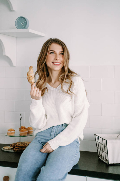 Beautiful blonde girl eating croissant in the morning in the kitchen - Photo, Image