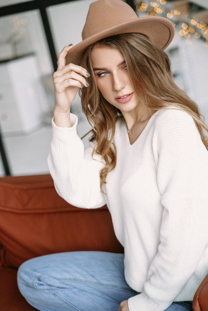 A beautiful blonde girl in a hat sits on a brown sofa in a stylish white apartment - Fotografie, Obrázek