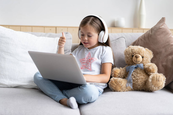 Online education. Sweet girl with headphones looking video lesson teacher conference laptop sitting on the couch at home - Фото, зображення