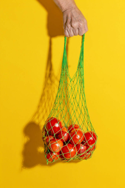 Male hand holding a green mesh bag with vegetables on yellow background - Photo, Image