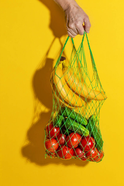 Erkek, sarı arka planda sebzeli beyaz bir ağ torbası tutuyor. - Fotoğraf, Görsel