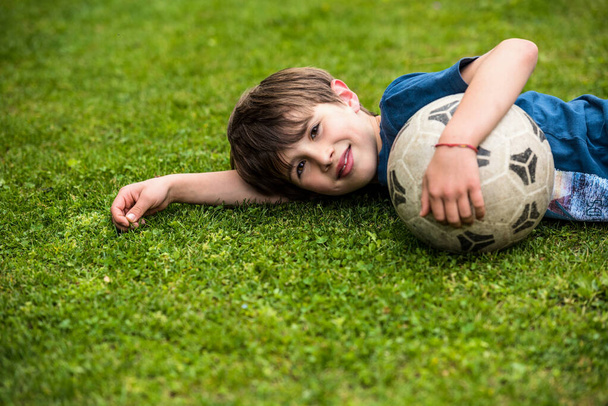 サッカーボールを持った子供が草の上に横になり、サッカーをした後に笑顔でリラックス - 写真・画像