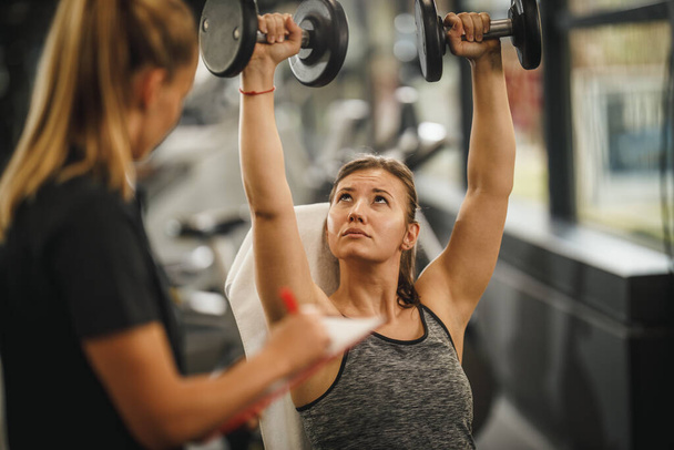 Una foto de una joven musculosa en ropa deportiva haciendo ejercicio con una entrenadora personal en el gimnasio. Ella está bombeando su músculo con mancuerna. - Foto, imagen