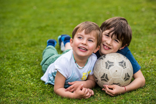 サッカーボールを持って芝生の上に寝そべっている2人の子供とサッカーをした後の笑顔でリラックス - 写真・画像