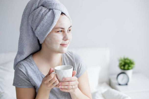 good morning and skin care concept - portrait of young woman with clay facial mask drinking coffee at home - 写真・画像