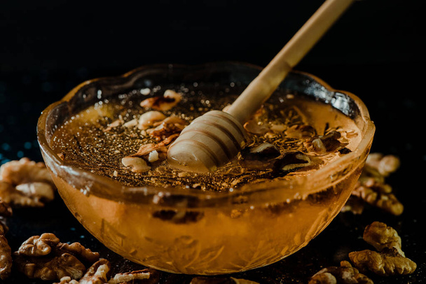 Wooden honey spoon dipped in a jar full of honey and nuts placed on a black background. Honey dripping around, pleasing and inviting photos. - Photo, image