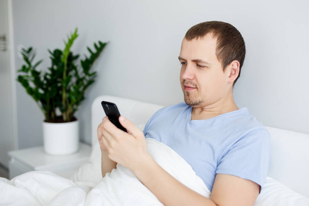 πορτρέτο του νεαρού όμορφου άνδρα που κάθεται στο κρεβάτι και χρησιμοποιεί smartphone - Φωτογραφία, εικόνα