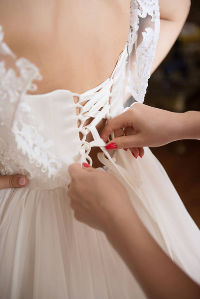 新婦付添人は結婚式の日のための花嫁を準備し、ボタンを助け、彼女の服を結ぶ。新婦付添人は花嫁の結婚式のドレス、花嫁の朝の集会の後ろに弓を作ります - 写真・画像