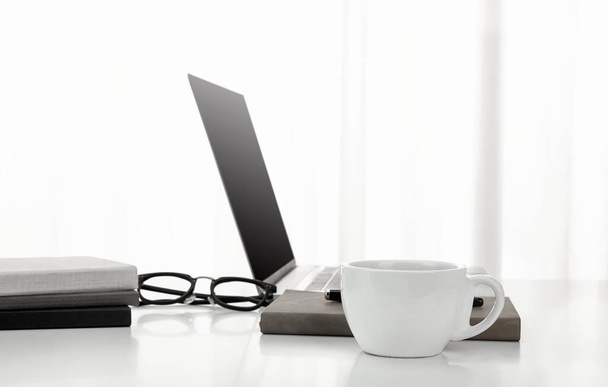 A closeup of a cup of coffee, laptop, notebooks, and a pair of glasses on a white desk - Foto, Imagem