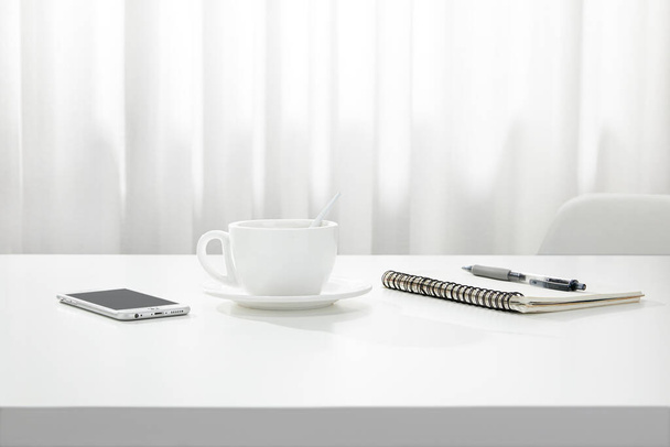 A closeup of a cup of coffee, a notebook and pen, and a smartphone on a white desk, indoors - Photo, Image
