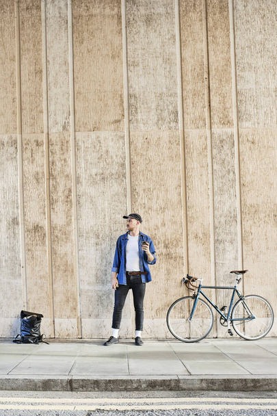 Full Length Of Professional Freelancer Young Man Standing With His Bike And Holding A Coffee Outside In Front Of A Wall. - Foto, afbeelding