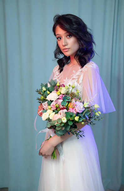 Портрет молодой невесты в модном свадебном платье с красивым макияжем и прической, студийное фото в помещении. Молодая, привлекательная модель азиатской внешности с цветами в руке - Фото, изображение