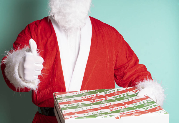 A closeup shot of Santa Claus holding a pizza box with thumbs up against a blue wall - Fotoğraf, Görsel