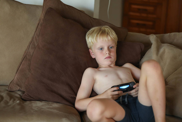 A young Caucasian boy playing video games at home on the sofa in soft afternoon light - Foto, Imagem