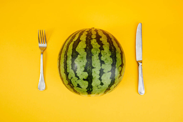 A watermelon with a fork and a knife next to it on a yellow background - Fotó, kép