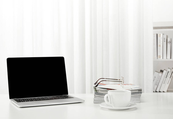 A closeup of a laptop, a cup of coffee, books, magazines, and more on a white desk, indoors - Photo, Image