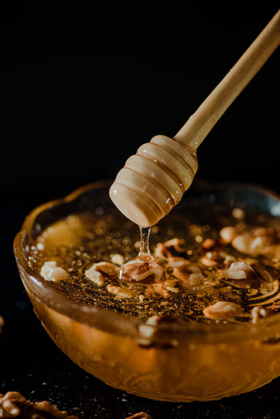 Wooden honey spoon dipped in a jar full of honey and nuts placed on a black background. Honey dripping around, pleasing and inviting photos. - Фото, изображение