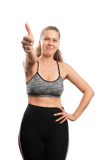 Fit weibliches Model lächelt in Sportbekleidung und zeigt Daumen hoch wie eine Geste des gesunden Lebensstils und Fitness-Konzepts auf weißem Hintergrund - Foto, Bild