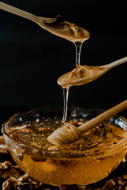 Wooden honey spoon dipped in a jar full of honey and nuts placed on a black background. Honey dripping around, pleasing and inviting photos. - Fotoğraf, Görsel