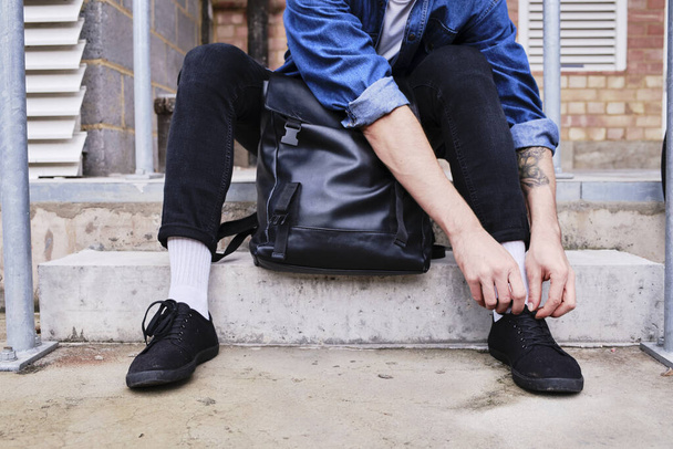 Young Man Sitting On The Street With His Bag Ties His Shoes On. - Foto, Bild