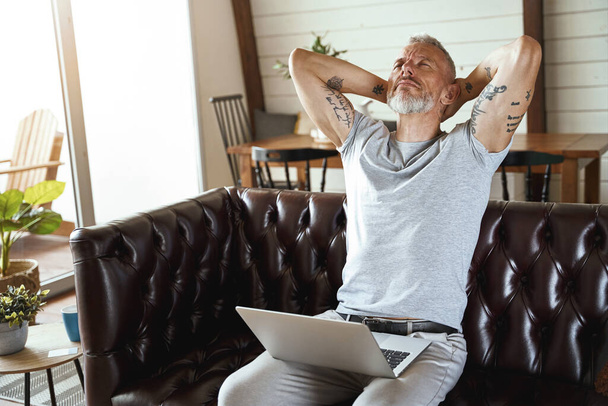 In search of inspiration. Thoughtful mature man in casual clothes looking upside and thinking about business while sitting on sofa in the living room, working on laptop - Photo, Image