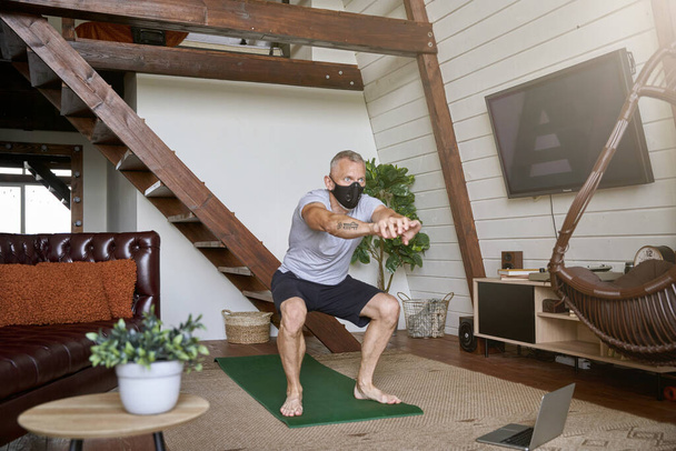 Оставаться активным в самоизоляции. Спортивный мужчина средних лет носит защитную маску для лица упражнения дома во время просмотра онлайн тренировки на ноутбуке - Фото, изображение