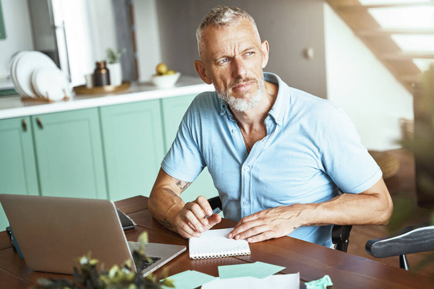 Домашний офис. Сосредоточенный белый мужчина средних лет делает заметки, сидя за столом и работая в Интернете - Фото, изображение