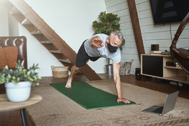 Middle aged man wearing face protective mask exercising at home in self isolation, standing in plank pose while watching online workout on laptop - Fotó, kép