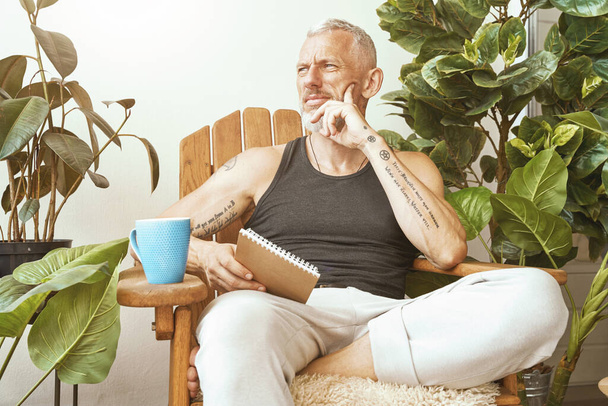 Kadonnut ajatuksiin. Keskittynyt keski-ikäinen mies istuu modernin asuntonsa aurinkoisella terassilla, pitäen muistikirjaansa ja miettien jotain. - Valokuva, kuva