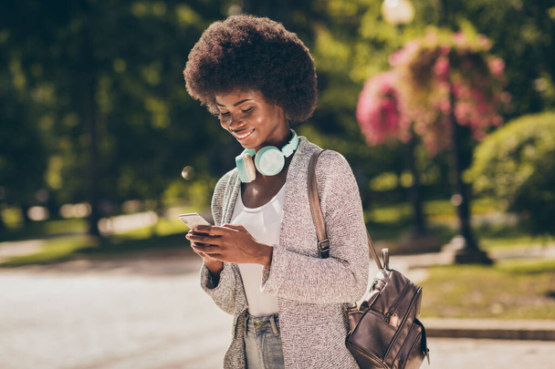 Photo of positive afro american girl use cellphone wear cardigan rucksack in town fall outside park - 写真・画像