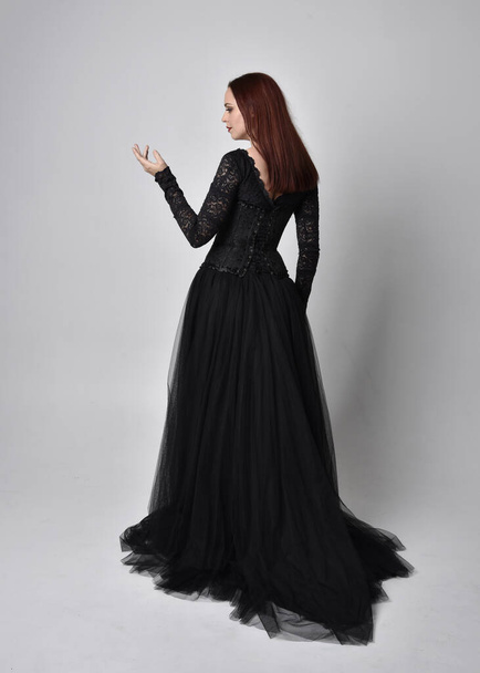Ganzkörperporträt einer Frau in schwarzem Gothikkleid, mit dem Rücken zur Kamera stehend vor Studiohintergrund. - Foto, Bild
