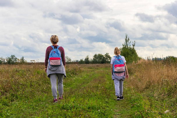Una madre y una hija caminan a lo largo de un camino que va muy lejos en el campo, a principios de otoño, sobre el fondo de un cielo con nubes gruesas. - Foto, imagen