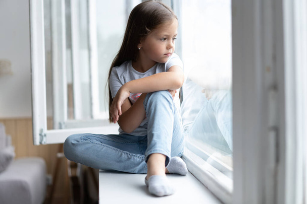 Beautiful little girl watching out the window. Young girl looking from window. Cheerful kid lies at windowsill - Фото, зображення