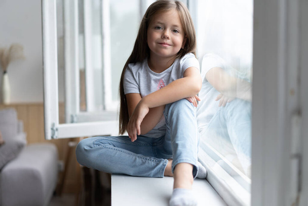 Beautiful little girl watching out the window. Young girl looking from window. Cheerful kid lies at windowsill - Fotoğraf, Görsel