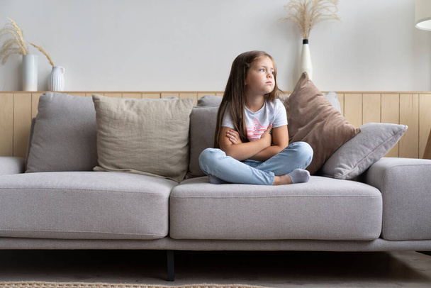 Beautiful unhappy sad little 6-year old girl sitting cross legged on couch in living room - Foto, Imagem